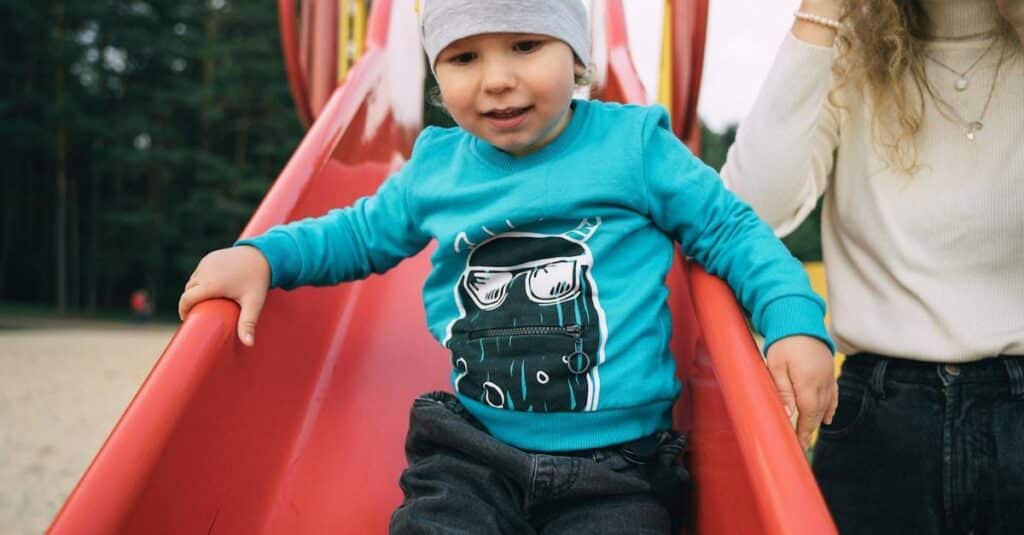 A toddler sliding down a red slide at a playground, enjoying a fun moment outdoors with a parent.