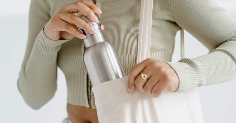 Close-up of woman placing a stainless steel water bottle into a cream-colored tote bag.