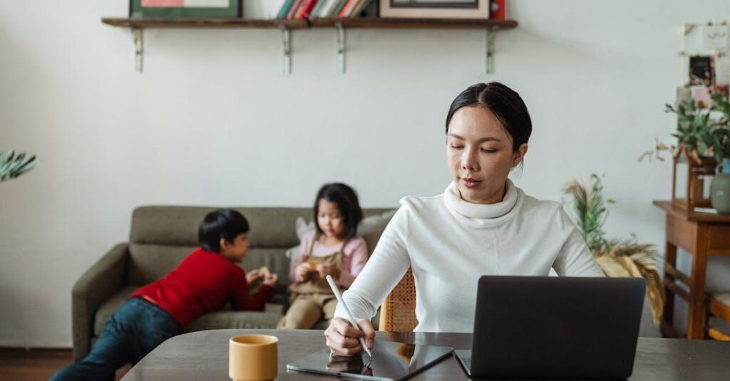 Mother multitasking with laptop and stylus alongside her children playing at home.