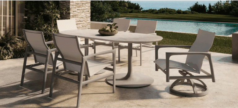 Luxury outdoor dining set with Castelle swivel rocker chair and sling-back chairs around an oval pedestal table on a stone patio overlooking a pool and ocean views.