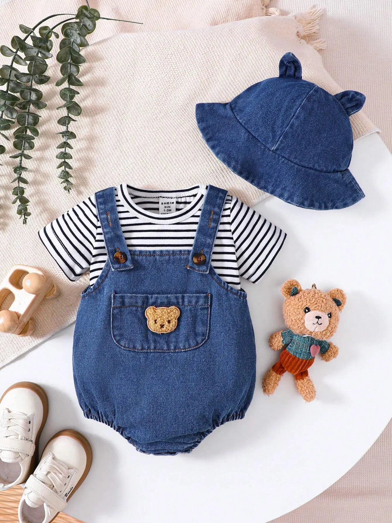Denim baby romper and matching hat on soft pink blanket with toy bear and baby shoes