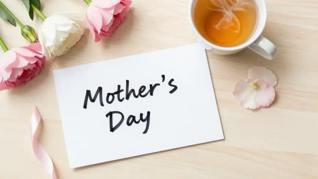 A Mother's Day scene featuring a handwritten card with “Mother's Day” on it, surrounded by soft pink and white flowers, a ribbon, and a cup of tea, creating a peaceful atmosphere.