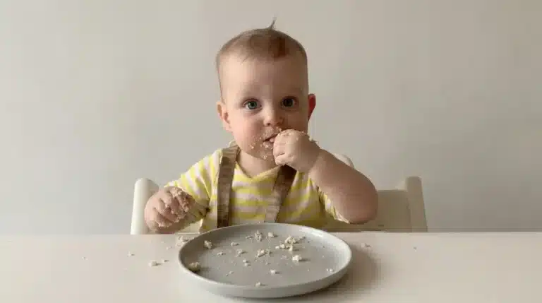 Baby-led weaning foods showing a baby eating finger foods independently at a table with small pieces of food on a plate and hands.