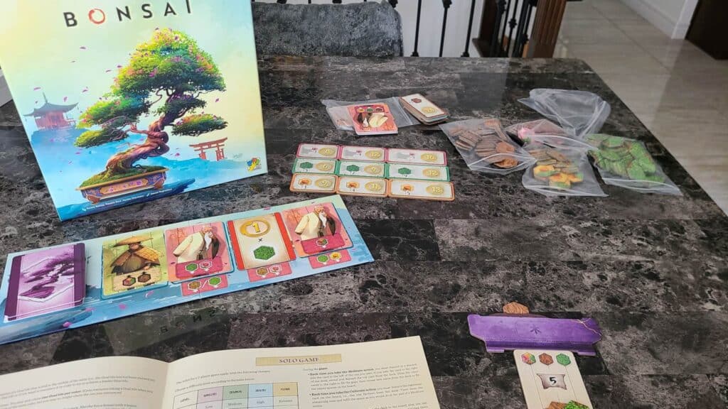 Bonsai board game components arranged on a table, including the illustrated game box, cards, tokens in plastic bags, player board, and rulebook