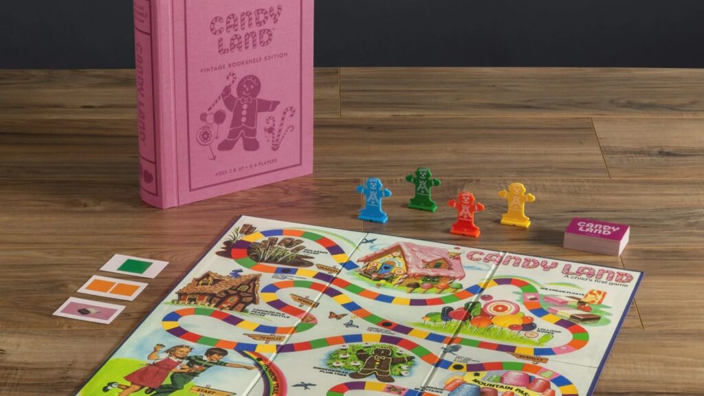 Candy Land board game set up on a wooden table, featuring the colorful game board, gingerbread player pieces in different colors, a deck of cards, and a pink vintage-style game box in the background