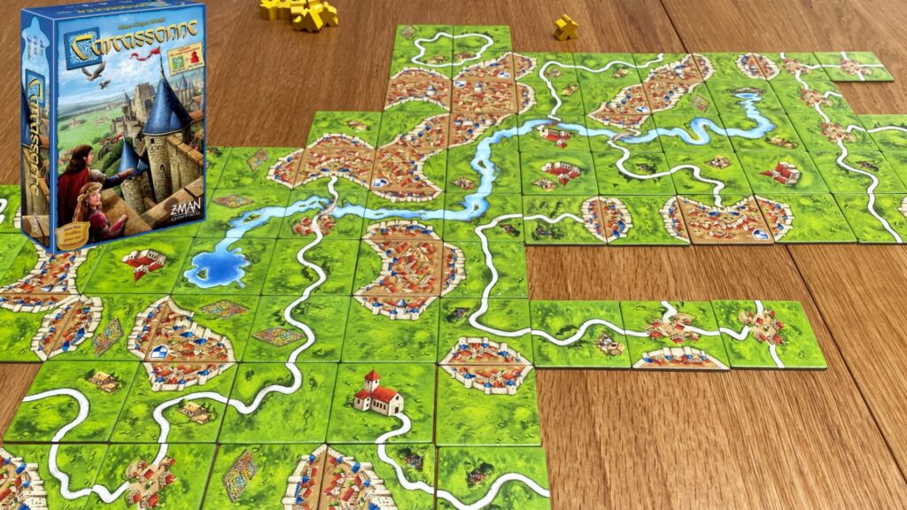 Carcassonne board game in progress with interlocking landscape tiles forming cities, roads, and rivers, with the game box placed beside the setup