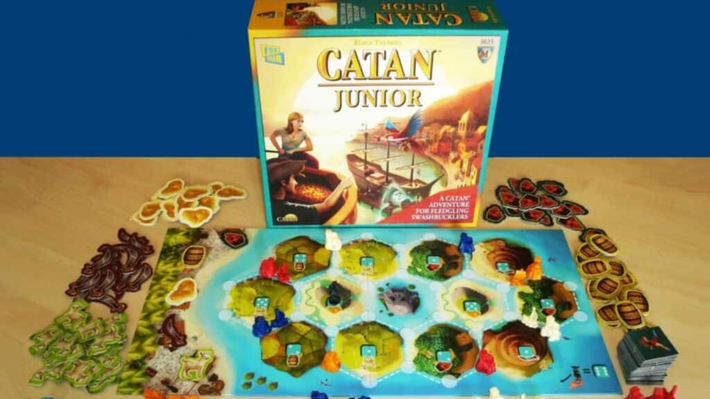 Catan Junior board game setup featuring the game board with island tiles, colorful player pieces, resource tokens, and the game box displayed upright in the background