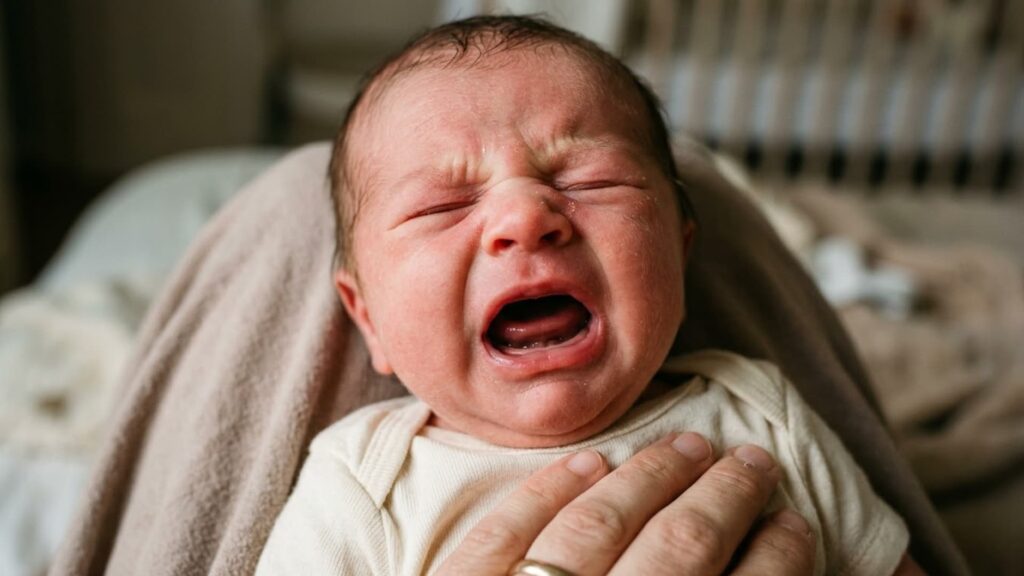 Close-up of a crying newborn baby being held, with eyes shut tightly and mouth open, showing signs of fussiness or discomfort.