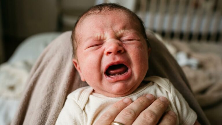 Close-up of a crying newborn baby being held, with eyes shut tightly and mouth open, showing signs of fussiness or discomfort.