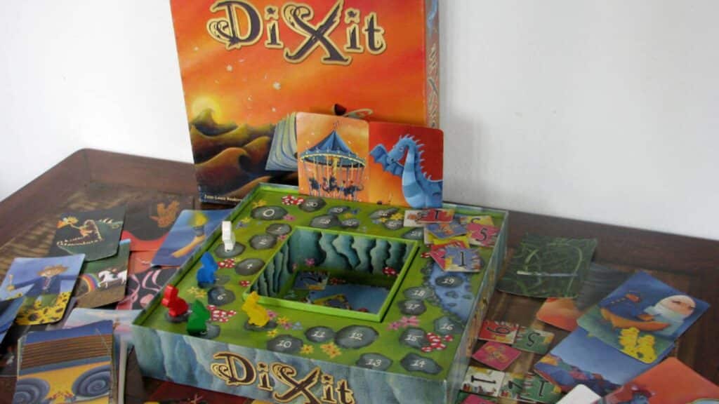 Dixit board game set up with illustrated cards, player pieces on a scoring track, and the game box displayed in the background
