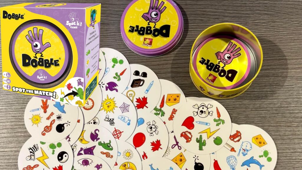 Dobble (Spot It!) card game set displayed on a wooden surface, featuring circular cards with colorful symbols spread out, along with the yellow tin case and branded game box