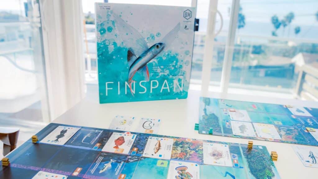Finspan board game setup on a table with ocean-themed player boards, cards, and tokens, with the game box featuring a flying fish displayed in the background near a window