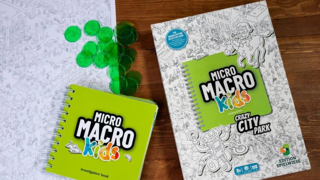 MicroMacro Kids board game materials displayed on a table, including a spiral-bound investigator’s book, a game booklet titled “Crazy City Park,” green tokens, and a black-and-white illustrated map