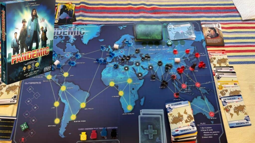 Pandemic board game setup showing a world map game board with colored disease cubes, player cards, tokens, and the game box displayed beside the board