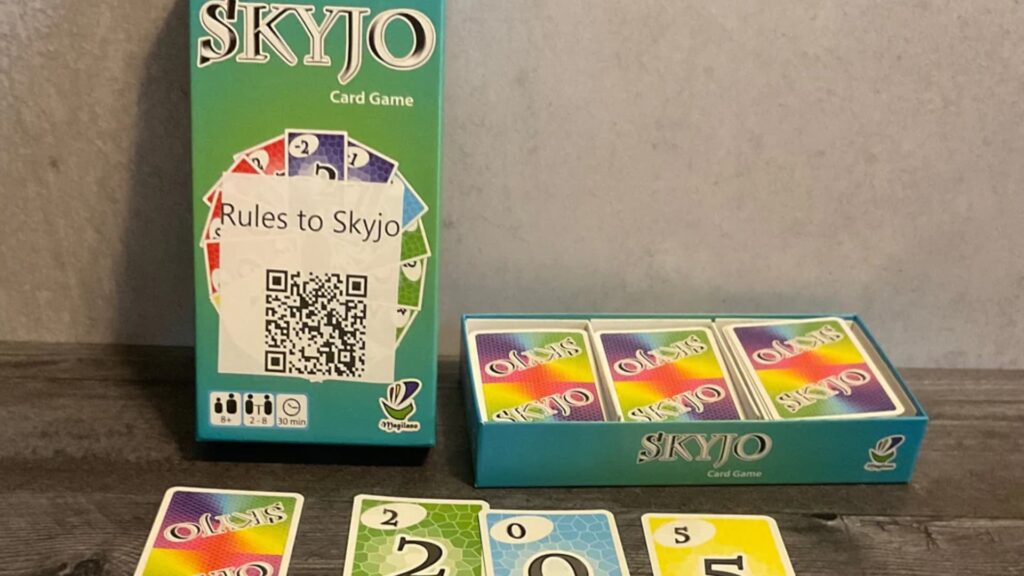 Skyjo card game setup showing the game box with a QR code for rules, an open tray with decks of cards, and several numbered cards laid out on a table