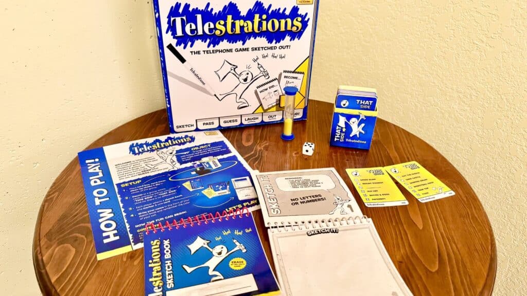 Telestrations party game components arranged on a wooden table, including the game box, sketchbooks, cards, dice, a timer, and an instruction sheet