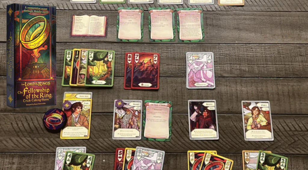 The Lord of the Rings Fellowship of the Ring trick-taking card game laid out on a wooden table, showing character cards, suit cards, and the game box alongside tokens and player hands