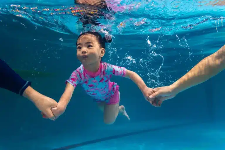 Child swimming underwater holding adult hands in a pool wearing pink swimwear