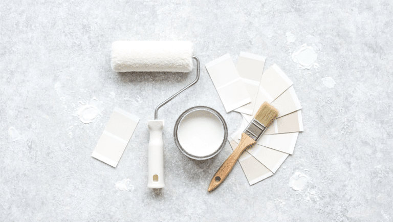 Paint roller, brush, open can of white paint, and neutral color swatches arranged on a textured surface.
