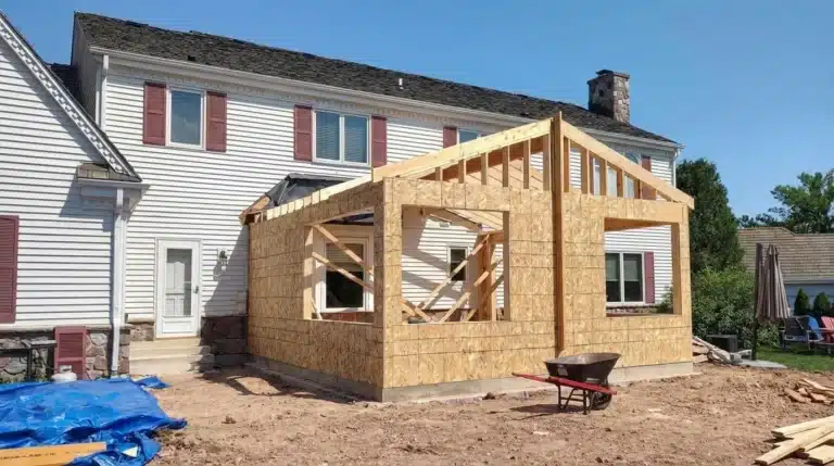 Home addition framing under construction behind a white house with exposed wood structure and tools nearby.