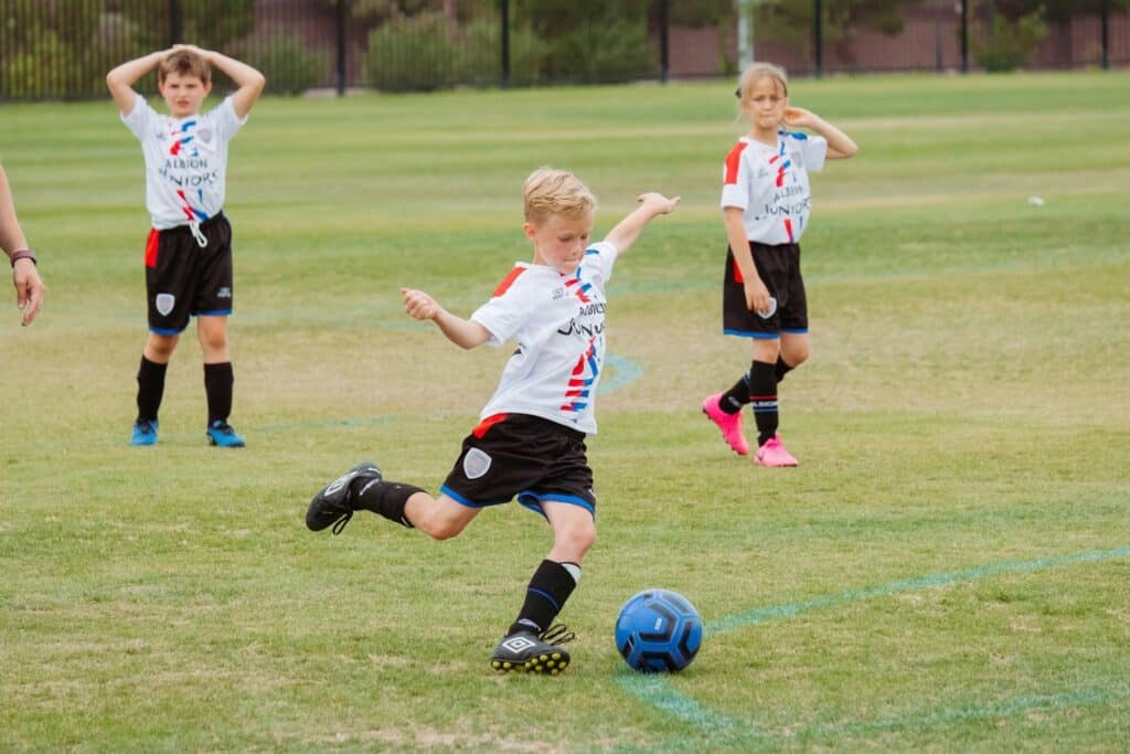 Child kicking a soccer ball on a grass field with teammates watching