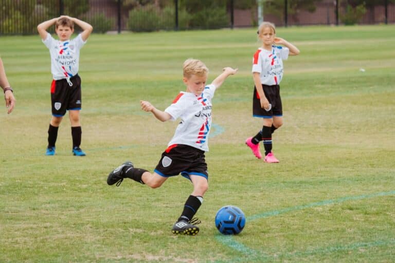 Child kicking a soccer ball on a grass field with teammates watching